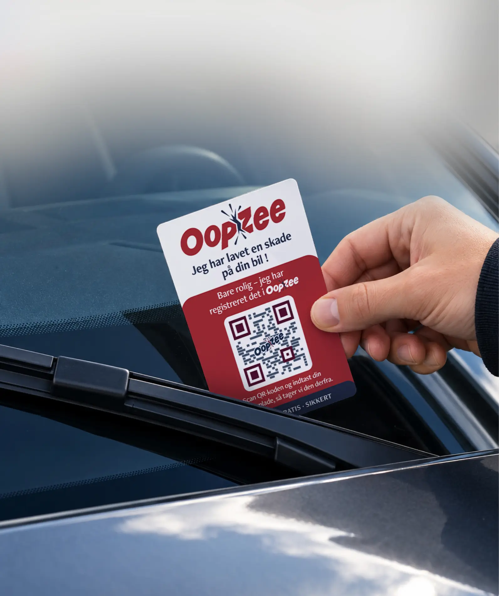 En hånd sætter et OopZee-kort med QR-kode under vinduesviskeren på en parkeret bil